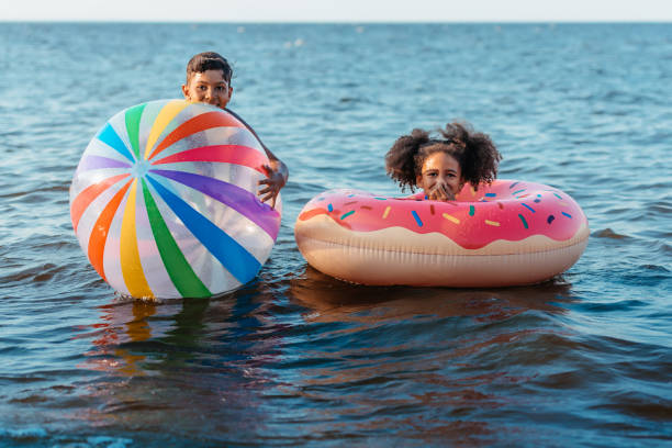 siblings cute smiling african american brother and sister having fun with beach ball and swimming tube in sea blow up doll stock pictures, royalty-free photos & images