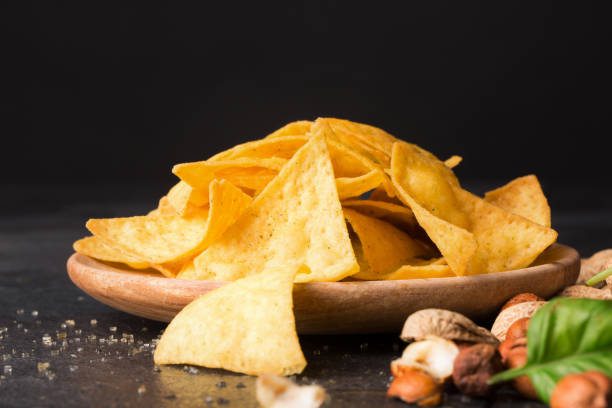 istock Close-up of crispy nachos on a a light wooden round plate. Corn chips next to nutsand basil leaves on a black background. 856571350