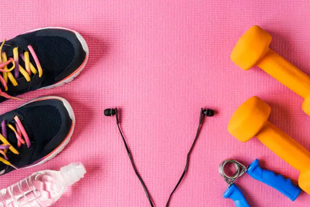 Sports accessories on a pink background. Sneakers, bottle of water, dumbbell, earphones. The place for the text.
