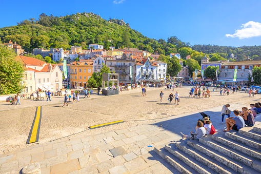 Sintra, Portugal - August 8, 2017: Tourists sitting on steps of National Palace of Sintra in the popular main Amelia Square with Castle of the Moors on background. Sintra urban center in a sunny day.