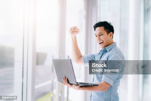 Foto de Animado Asiático Homem Segurando Laptop E Levantando O Braço Para Comemorar O Sucesso e mais fotos de stock de Homens
