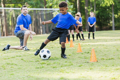 A 7 year old African-American boy on a soccer team at practicing, kicking a ball around cones. The coach is watching and other children are waiting their turn to do the drill.