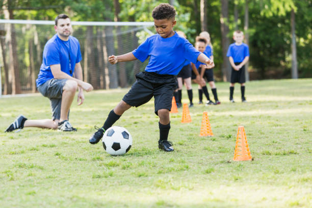 Boy on soccer team practicing A 7 year old African-American boy on a soccer team at practicing, kicking a ball around cones. The coach is watching and other children are waiting their turn to do the drill. soccer soccer ball kicking adult stock pictures, royalty-free photos & images