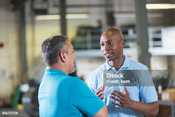 Two Multiethnic Workers In A Workshop Talking Stock Photo - Download Image Now - Talking, Blue-collar Worker, Warehouse