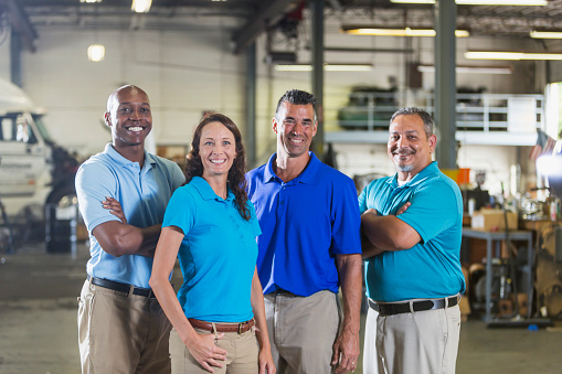 A multi-ethnic group of four workers, including a woman, working for a trucking company. They are smiling at the camera. A semi-truck with an open hood is out of focus in the background.