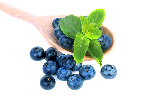 Close-up of a pile of sweet blueberries and green mint leaves in a wooden spoon, isolated on a white background. Some berries near the spoon with nutritious bilberries. Copy space.