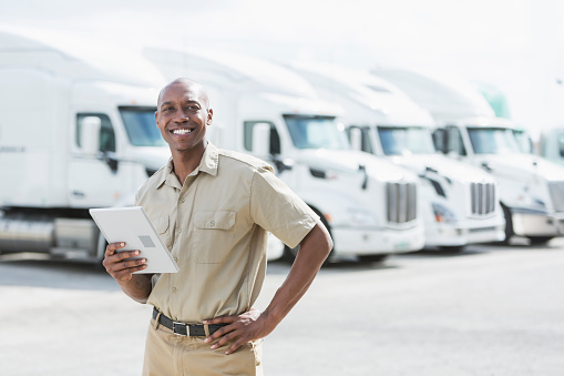 An African-American man in his 30s standing in front of a row of parked semi-trucks outside a distribution warehouse, holding a digital tablet.