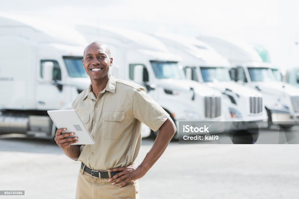 Black man standing in front of semi-trucks An African-American man in his 30s standing in front of a row of parked semi-trucks outside a distribution warehouse, holding a digital tablet. Fleet of Vehicles Stock Photo