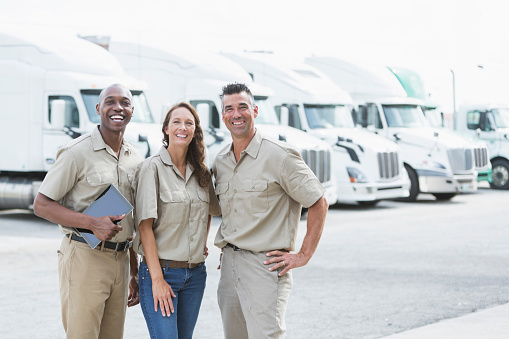 A group of three multi-ethnic workers standing in front of a row of parked semi-trucks at a distribution warehouse. The woman standing in the middle and the Caucasian man are in their 40s. The African American man is in his 30s.