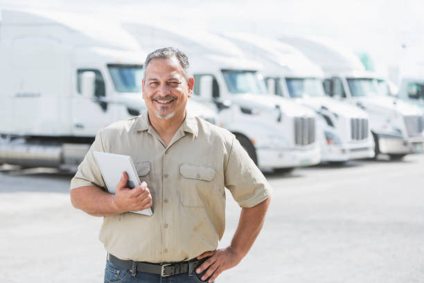 Hispanic man standing in front of semi-trucks An Hispanic man in his 40s standing in front of a row of parked semi-trucks outside a distribution warehouse, holding a digital tablet. driver occupation photos stock pictures, royalty-free photos & images