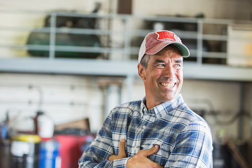 A mature man in his 40s standing in a garage or warehouse, wearing a trucker's hat and plaid shirt, arms crossed, smiling.