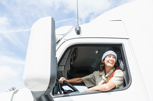 A mature woman driving a semi-truck. She is smiling, looking out of the driver's side window over her shoulder.