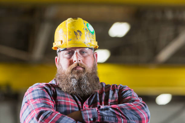 Bearded construction worker wearing hardhat A serious man, a construction worker with a long beard wearing a hardhat, safety glasses and a plaid shirt. He is looking at the camera with his arms crossed man beard plaid shirt stock pictures, royalty-free photos & images