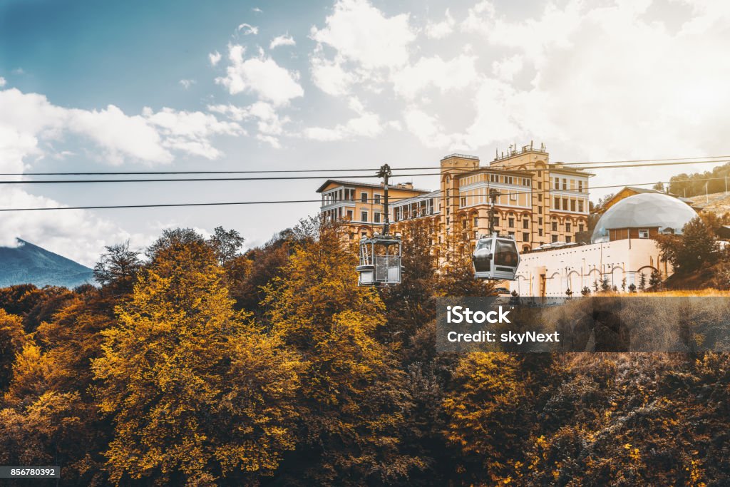 Cableway during bright autumn day Autumn landscape in resort district of Sochi city, Russia: yellow trees, ropeway above them with one modern funicular cabin and retro one, dry grass; resort buildings and hills in background Autumn Stock Photo