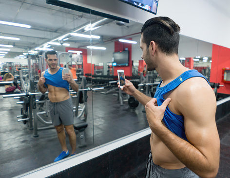 Happy man taking a selfie at the gym with a cell phone showing off his muscles â sports training concepts
