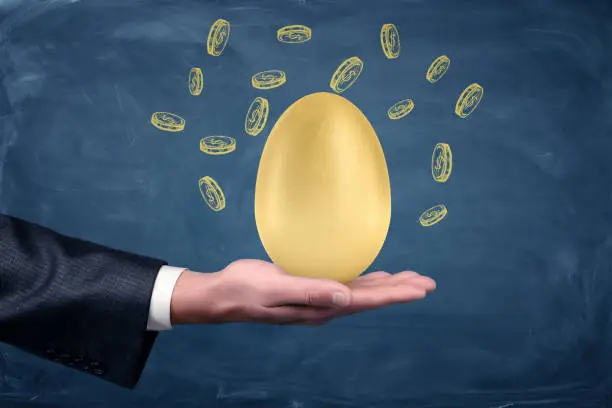 Photo of A businessman's hand holding a large golden egg on a blue blackboard background with drawn dollar coins.