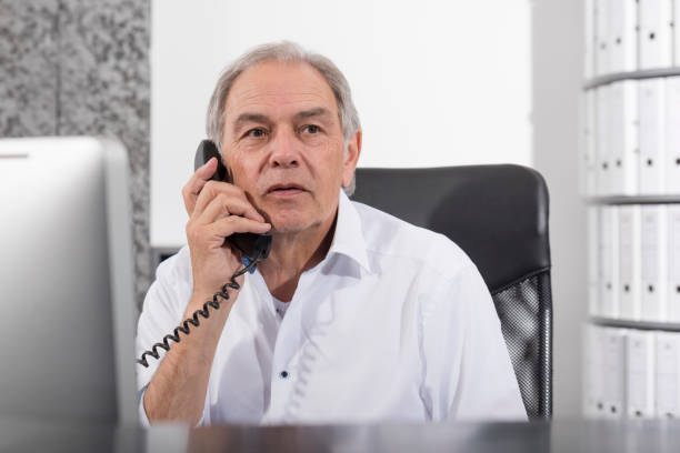 Telephoning older man with white shirt A middle-aged man in the office who speaks into a telephone. With a computer on his desk and a filing cabinet in the background.ender older man with a white shirt sits at a desk büro stock pictures, royalty-free photos & images