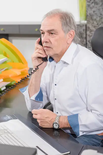Elderly man on the phone with a white shirt sits at a desk