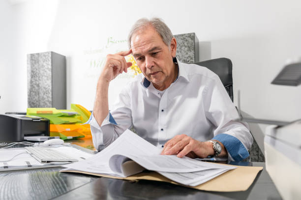 Older man with white shirt sits at a desk and leafs through a document file Older man with a white shirt sits at a desk and leafs through a file in a document fähigkeit stock pictures, royalty-free photos & images