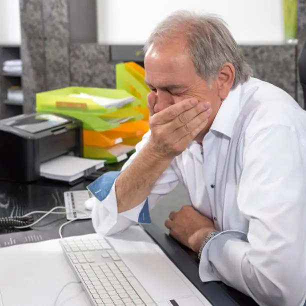 Older man sits desperately at a desk with his hand in front of his mouth