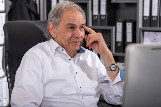 Smiling older man with white shirt Smiling older man with a white shirt sits at a desk fähigkeit stock pictures, royalty-free photos & images