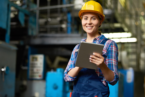 Portrait of female factory worker using digital tablet and smiling, looking at camera posing in modern workshop