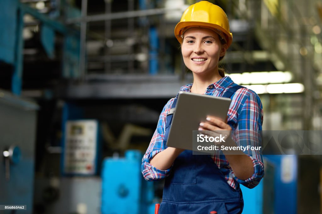Beautiful Young Woman Working at Modern Plant Portrait of female factory worker using digital tablet and smiling, looking at camera posing in modern workshop Manufacturing Occupation Stock Photo