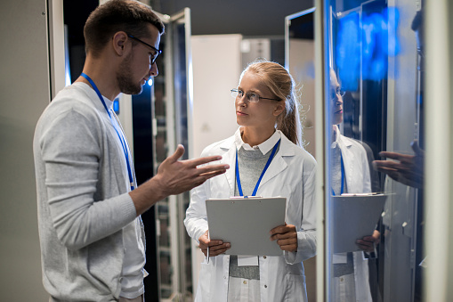 Portrait of  two scientists, man and woman, standing by server cabinets and discussing data while working with supercomputer in research center