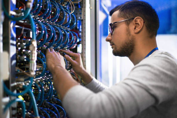 System Administrator Checking Servers Side  view portrait of young man connecting cables in server cabinet while working with supercomputer in data center connecters stock pictures, royalty-free photos & images