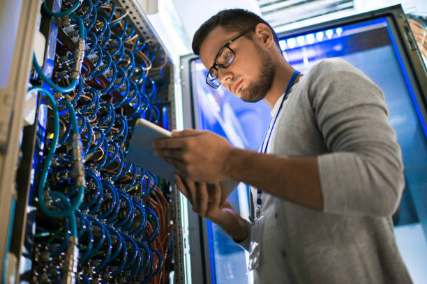 Young Network Engineer Low angle portrait of young man using digital tablet standing by server cabinet while working with supercomputer in blue light supercomputer stock pictures, royalty-free photos & images