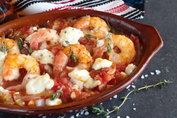 Prawns in a tomato sauce with feta cheese, garlic in a tradtitional greek ceramic bowl with fresh thyme, seasalt on a black abstract background. Greek healthy food.