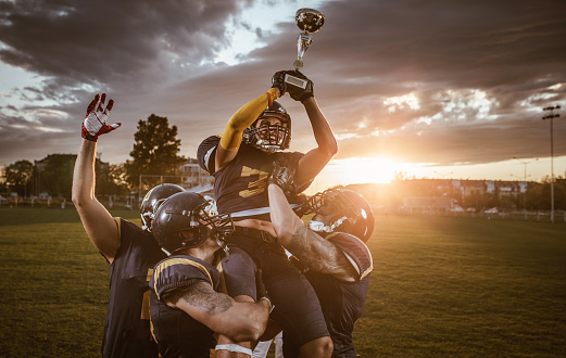 American football players holding their teammate high up who is holding a trophy while celebrating a victory.