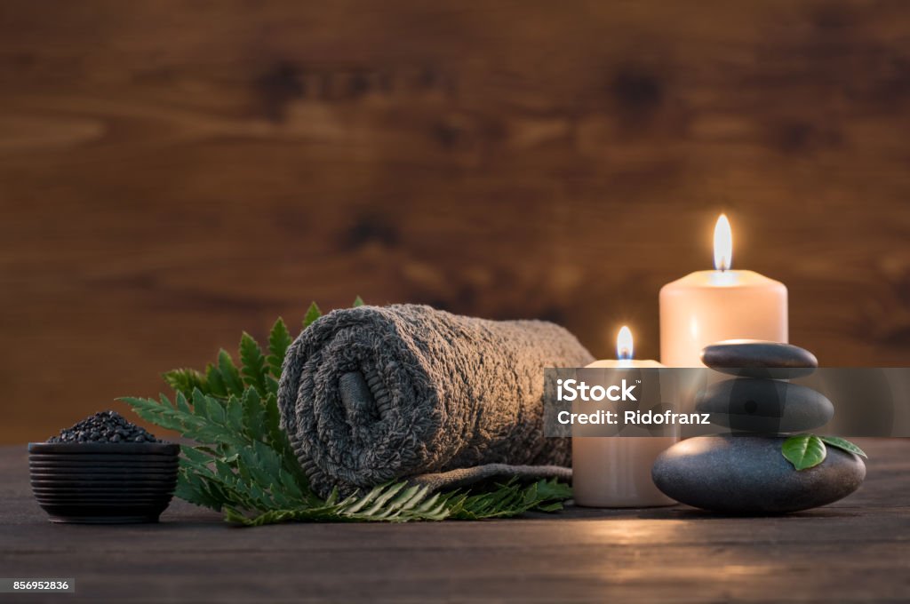 Spa treatment set Brown towel on green fern with candles and black hot stone on wooden background. Hot stone massage setting lit by candles. Hot stone therapy for one person with candle light. Beauty spa treatment and relax concept. Spa Stock Photo