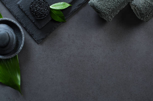 Luxury black spa Top view of two gray rolled towels with hot stones pile with copy space. High angle view of male beauty treatment set with green leaf and black salt. Luxury and elegant spa setting on black background. black male massage stock pictures, royalty-free photos & images