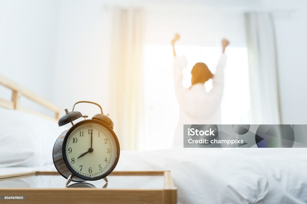Closeup alarm clock, Young asian woman wake up in the moring and stiing on bed at mirror door side relaxing in holiday with sunlight Sleeping Stock Photo