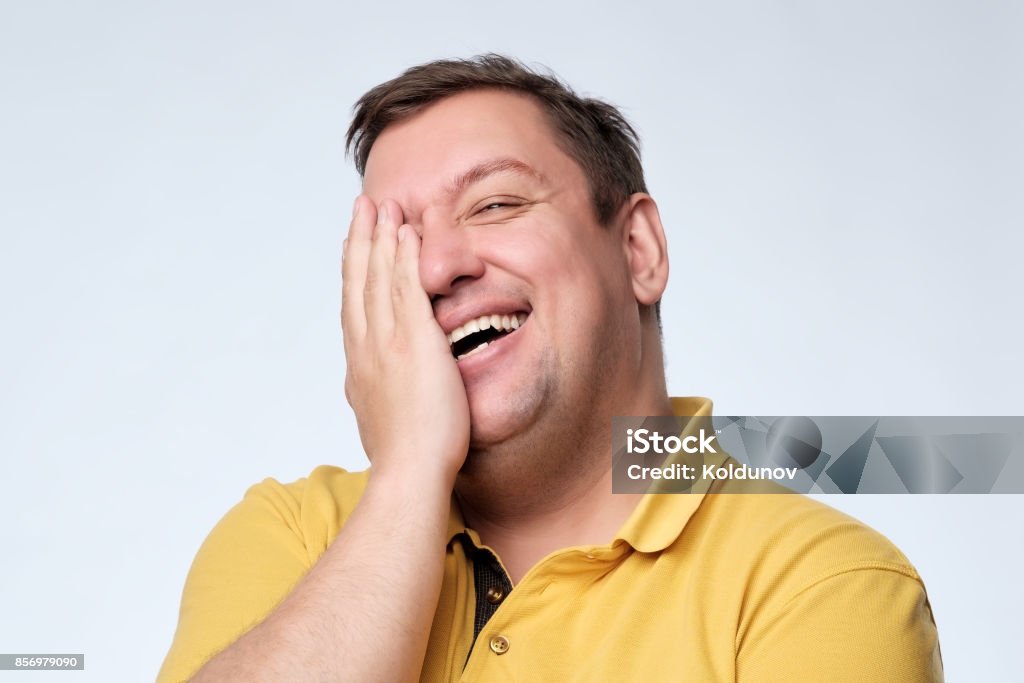 Caucasian fat man laughs and covers his face with his hands. Caucasian fat man in yellow t-shirt laughs and covers his face with his hands. Positive facial expression. Laughing Stock Photo