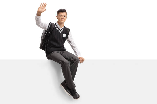 Teenage student in a uniform sitting on a panel and waving at the camera isolated on white background