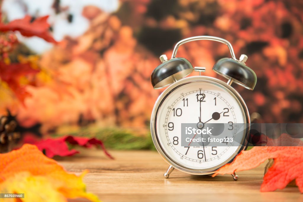 Daylight savings time.  2 o'clock time on clock in autumn season. It's daylight savings time.  The alarm clock is set for 2 o'clock am on an outdoor table with autumn trees, leaves in background.  Copyspace at side. Daylight Saving Time Stock Photo