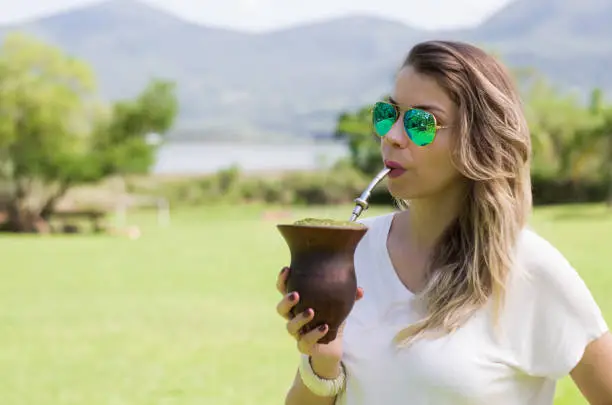 Blonde woman drinking the traditional Chimarrão from the State of Rio Grande do Sul in Brazil. Culture Gaúcha, Brazil. A type of Mate herb tea (Ilex paraguariensis) served in the cuia (fruit of the breeding ground (Lagenaria siceraria), drink with a kind of stainless steel straw (pump).