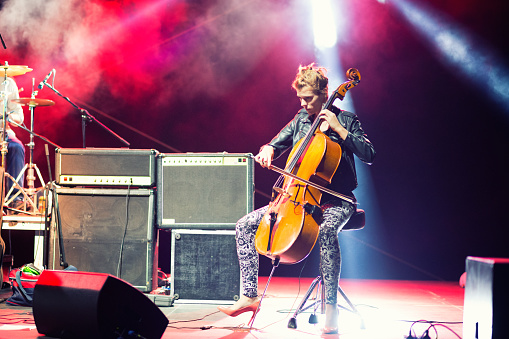 Young female musician playing cello on stage as a member of rock and roll band in concert.