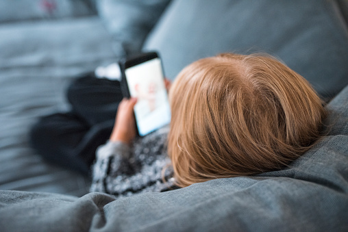 Young blond girl is sitting on the sofa and using mobile phone.