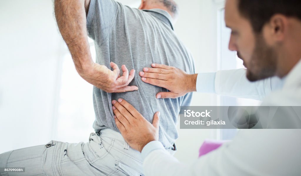 Back problems. Closeup rear low angle view of an early 60's senior gentleman having some back pain. He's at doctor's office having medical examination by a male doctor. The patient is pointing to his lumbar region. Backache Stock Photo