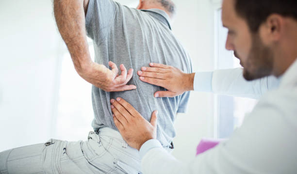 Back problems. Closeup rear low angle view of an early 60's senior gentleman having some back pain. He's at doctor's office having medical examination by a male doctor. The patient is pointing to his lumbar region. human spine stock pictures, royalty-free photos & images