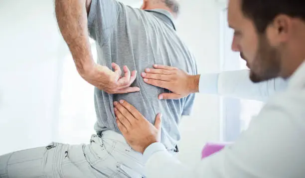 Closeup rear low angle view of an early 60's senior gentleman having some back pain. He's at doctor's office having medical examination by a male doctor. The patient is pointing to his lumbar region.