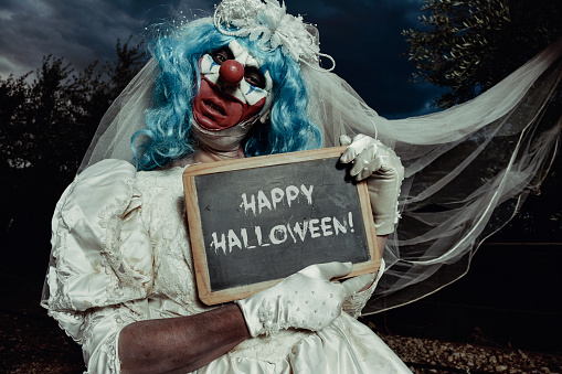 a scary evil clown wearing a dirty and ragged bride dress in a disturbing rural landscape at night holds a rustic chalkboard with the text happy halloween written in it