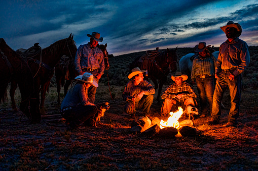 Cowboys and Cowgirls gather around a campfire in an open range with their horses and dog