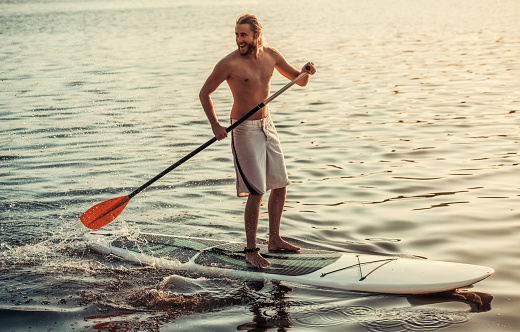 Handsome young man is smiling while SUP surfing on the river