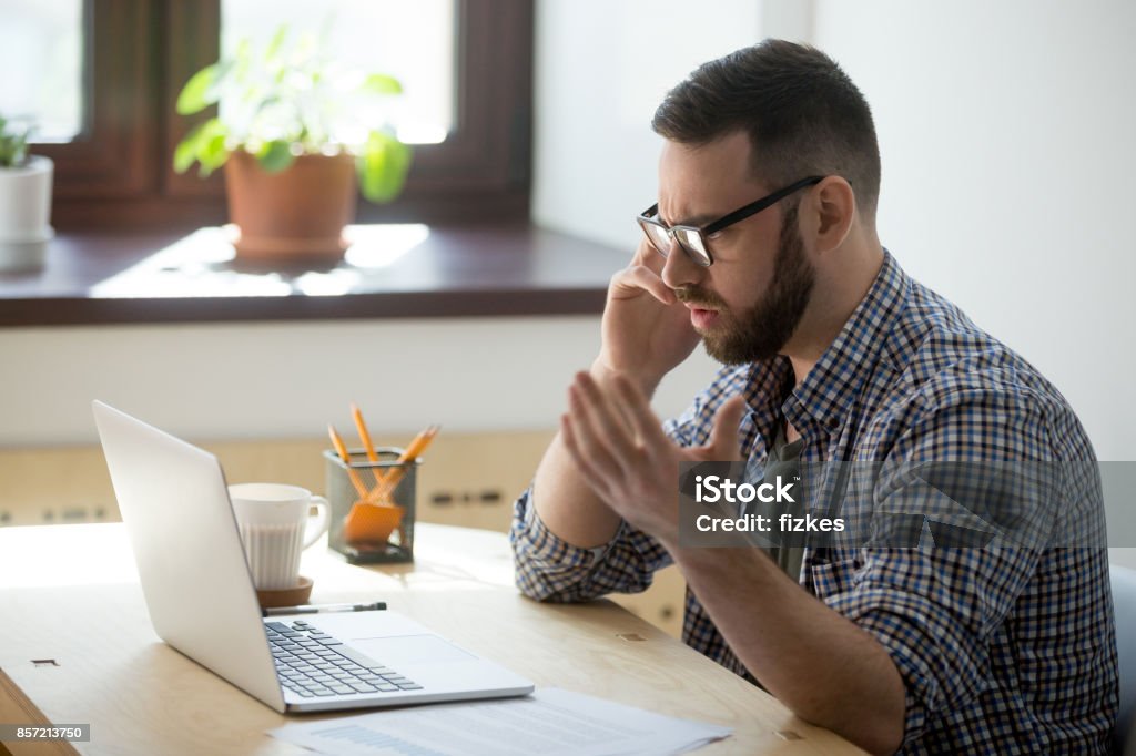 Frustrated male employee discussing contract details over the phone. Frustrated male employee discussing contract details over the phone. He is looking at documents in his laptop, holding phone, fling arms in an angry gesture, trying to find solution. Customer Stock Photo