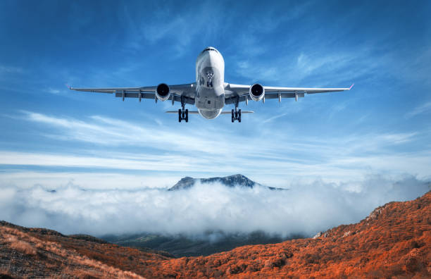 Airplane is flying over low clouds and mountains with autumn forest. Amazing landscape with passenger airplane, trees, mountains, blue cloudy sky. Passenger aircraft. Business travel. Commercial plane stock photo