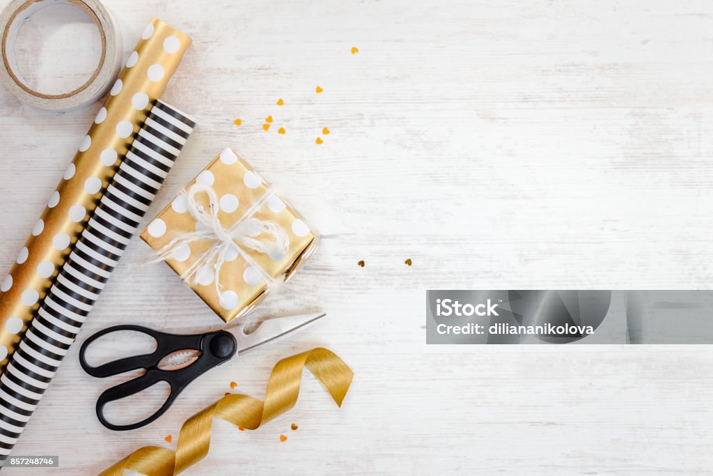 Gift box wrapped in golden dotted paper and wrapping materials on a white wood old background. Empty space. Wrapping Paper Stock Photo